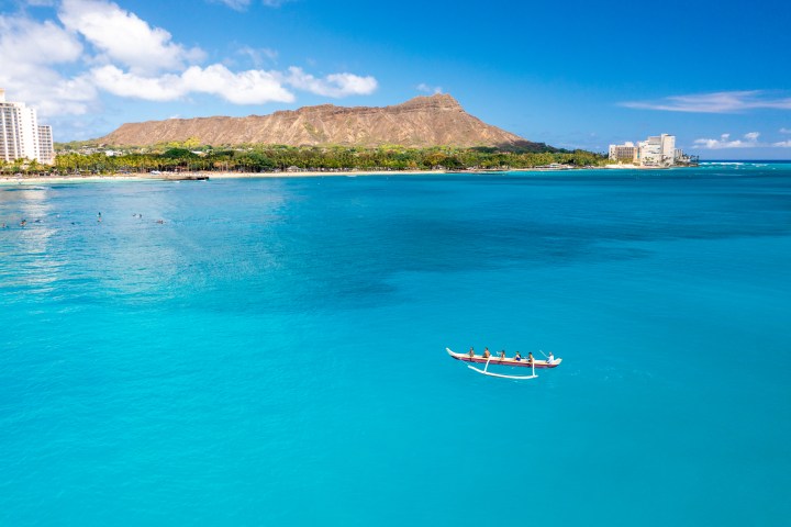 A canoe with four people on turquoise waters near a beach with a mountain in the background.