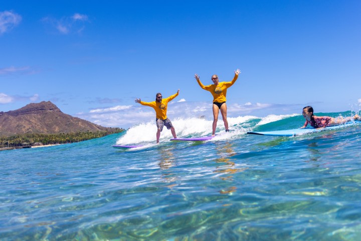 Three people surfing on clear, blue ocean waves under a bright sky near a mountainous coastline.