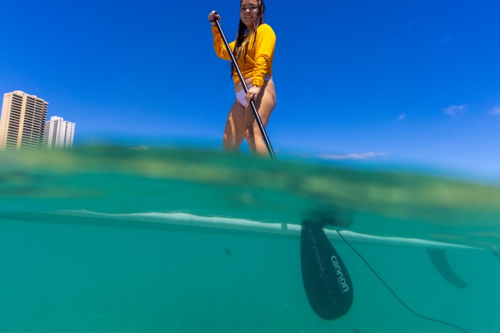 Woman paddleboarding on clear water with buildings in the background.